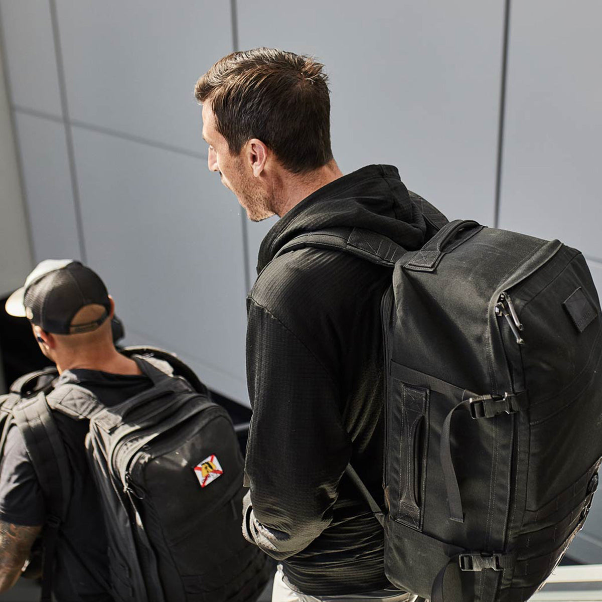 Two men with black backpacks walk down a staircase in a modern, well-lit indoor setting.
