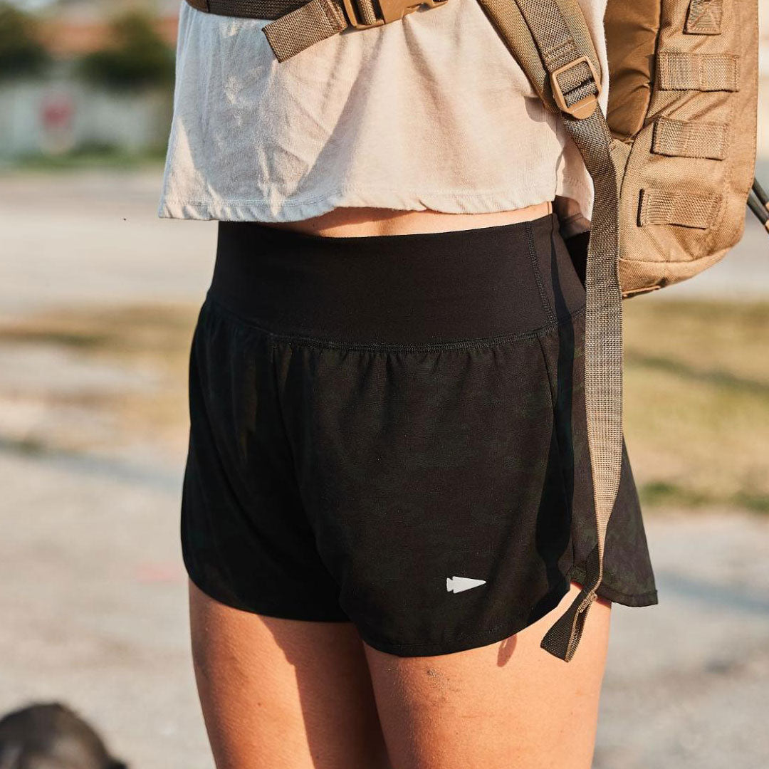 Person wearing black athletic shorts and a tan backpack, standing outdoors in sunlight.