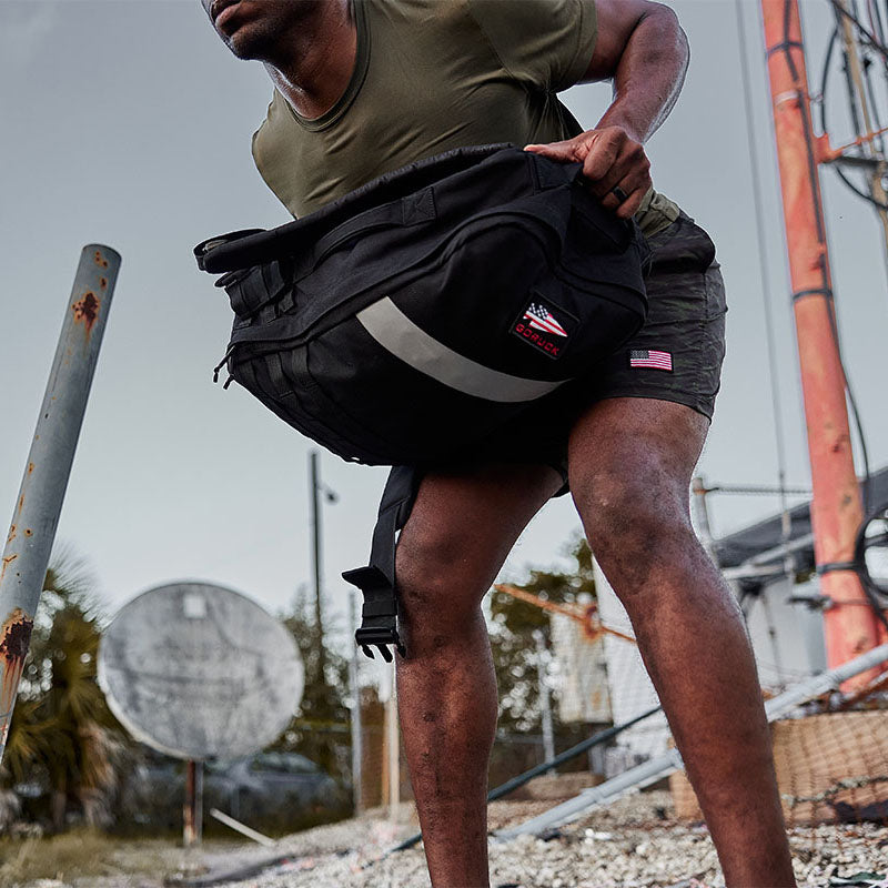 A person in shorts and a t-shirt bends down, holding a black bag outdoors near a satellite dish.