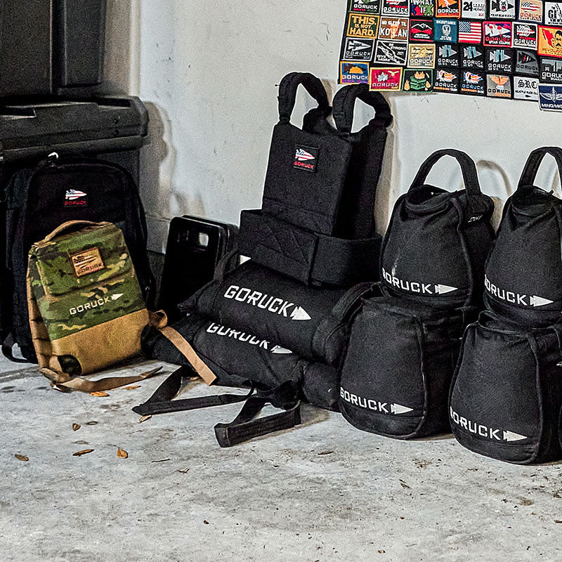 GORUCK backpacks and gym bags stacked on a concrete floor near a wall covered with colorful patch badges.