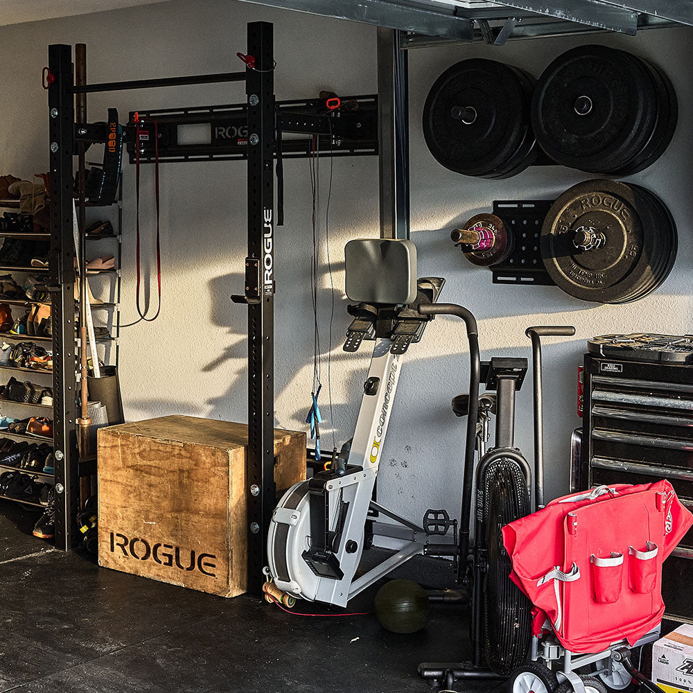 Home gym with a squat rack, weights, exercise bike, wooden box, and a red stroller in the foreground.