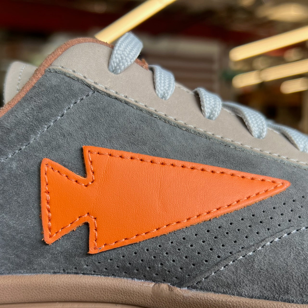 Close-up of the Mackall 45 Fog sneaker featuring gray suede, white laces, and an orange arrow-shaped patch on the side.