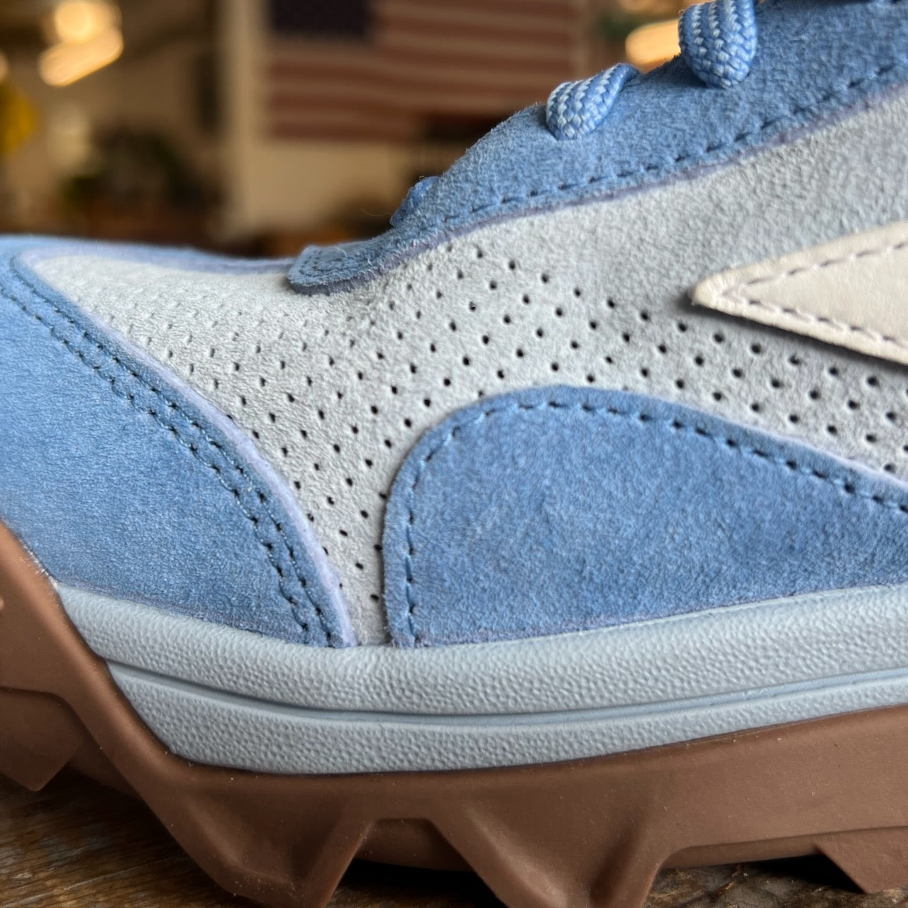 Close-up of the Mackall 45 Americana sneaker in blue and gray with a brown sole, displayed indoors against a blurred American flag backdrop.