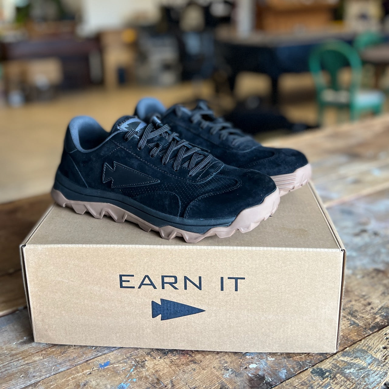 The Mackall 45 Black + Gum sneakers, featuring tan soles, displayed on a cardboard box labeled “EARN IT” with an arrow symbol.