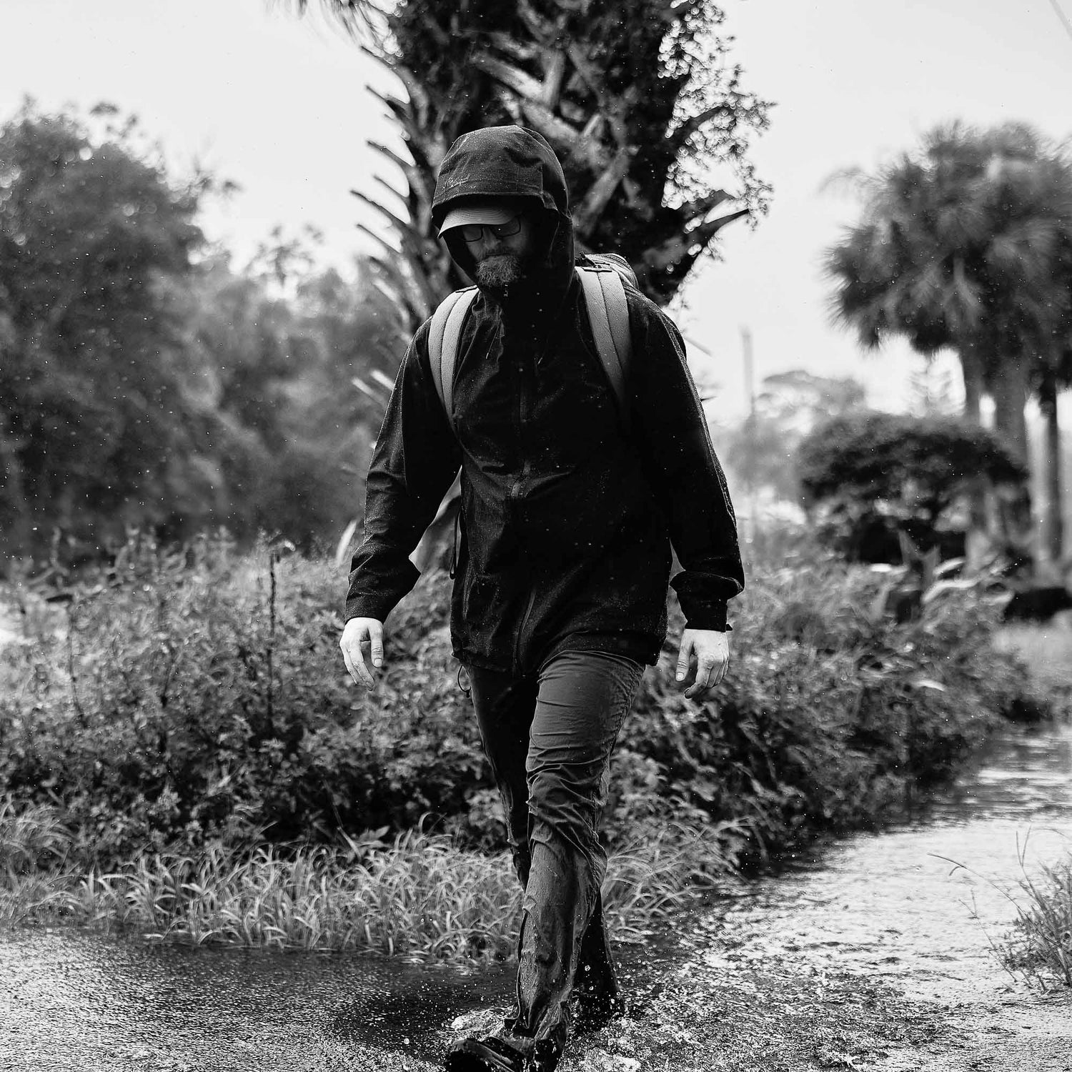 A person in a hooded jacket and backpack walks through a puddle on a rainy day.