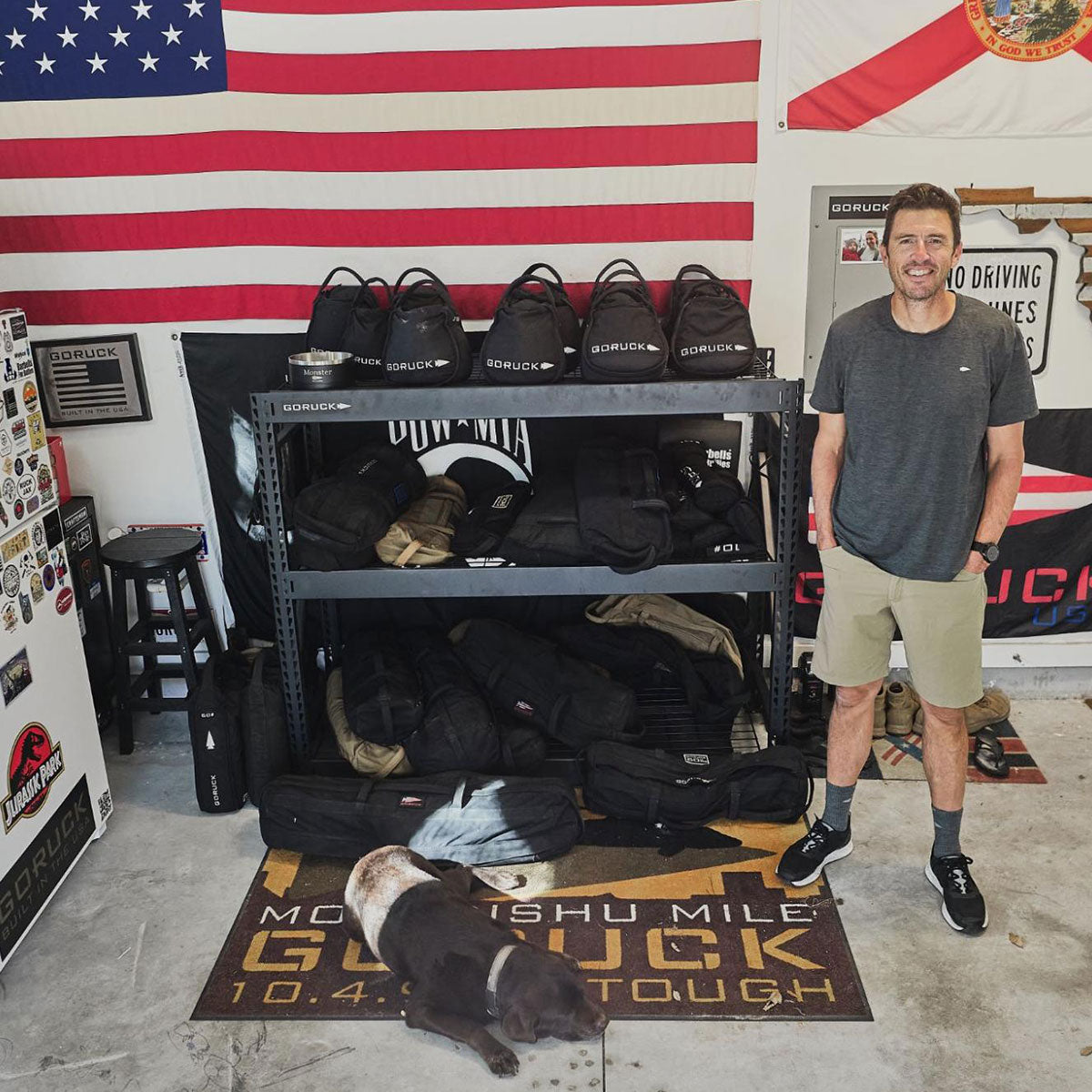 A man stands smiling by shelves of GORUCK gear, with a dog lying on a GORUCK mat and flags on the wall.
