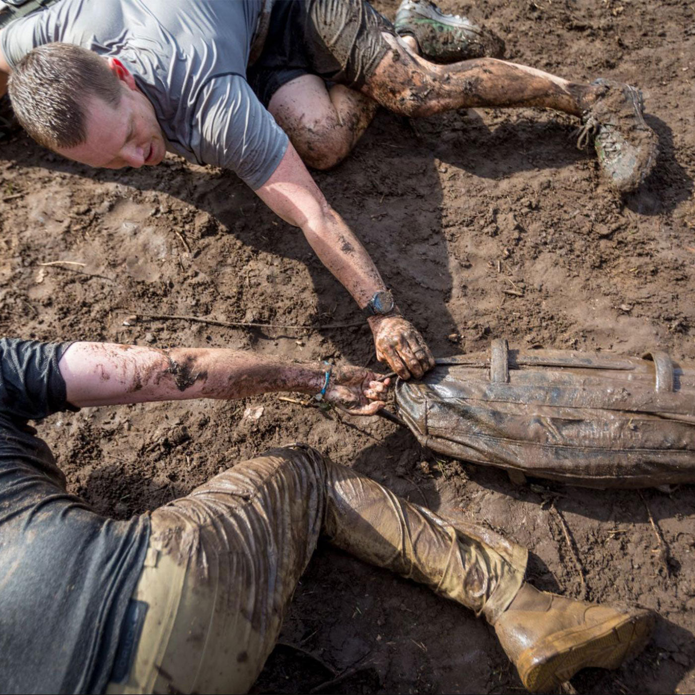 Two people covered in mud reach for a muddy backpack while lying on a dirt ground.