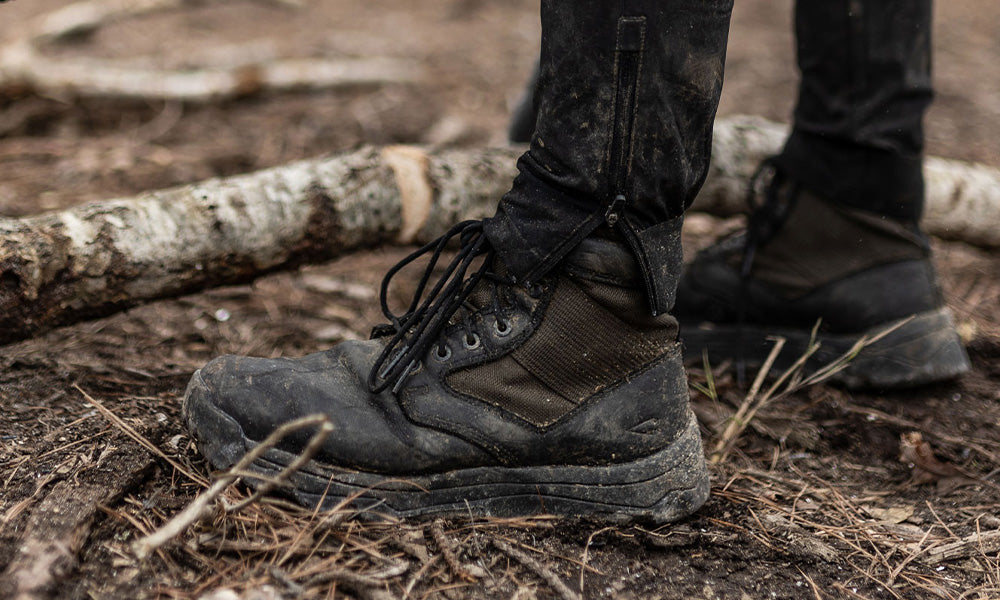 Close-up of muddy black boots and pants standing on dirt and fallen branches in an outdoor setting.