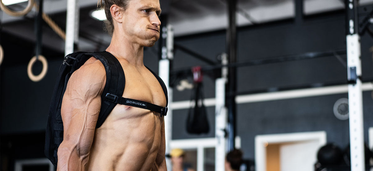 Muscular man with a backpack lifts weights in a gym, cheeks puffed, veins visible on his arms and chest.