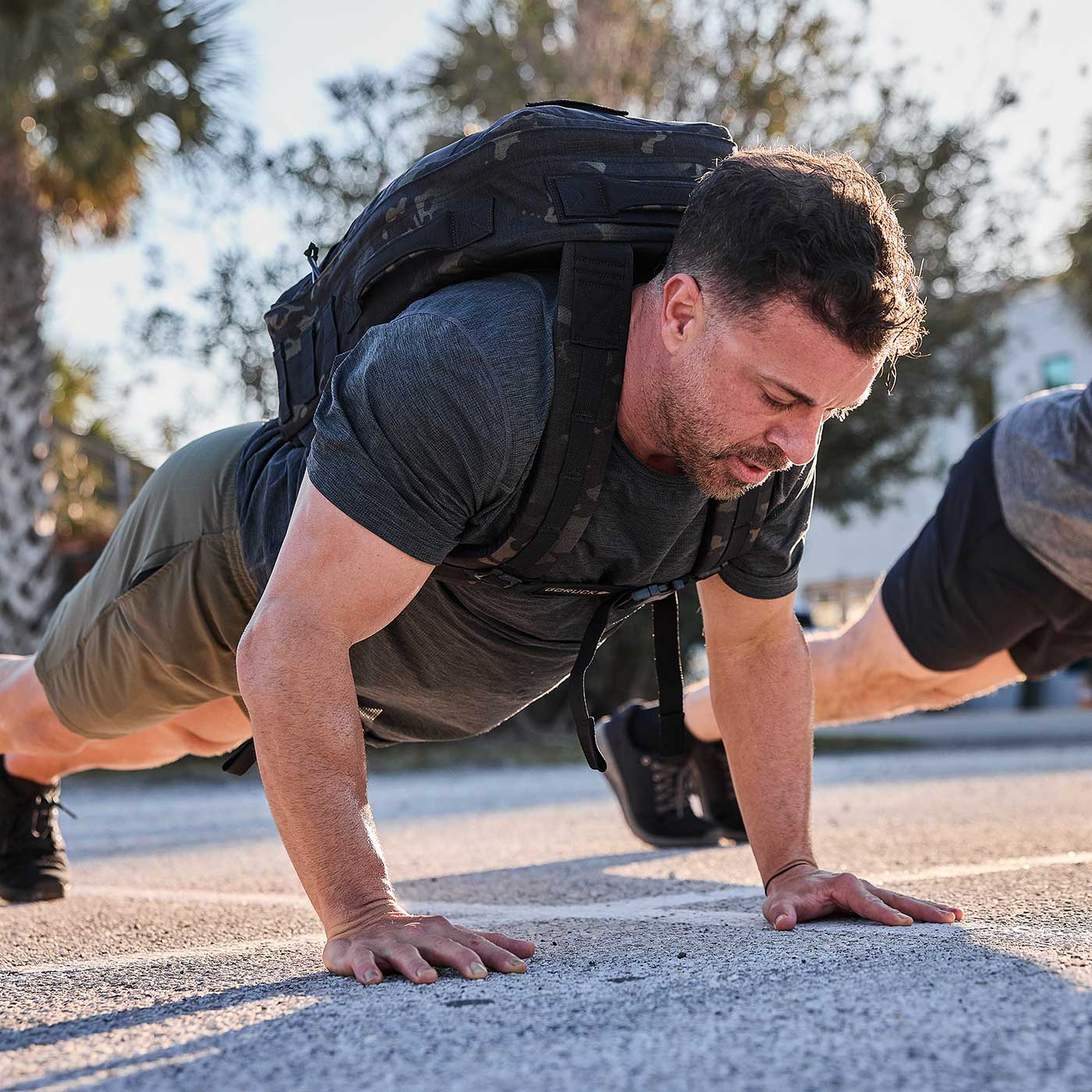 Man doing push-ups outdoors while wearing a backpack, with sunlight and trees in the background.