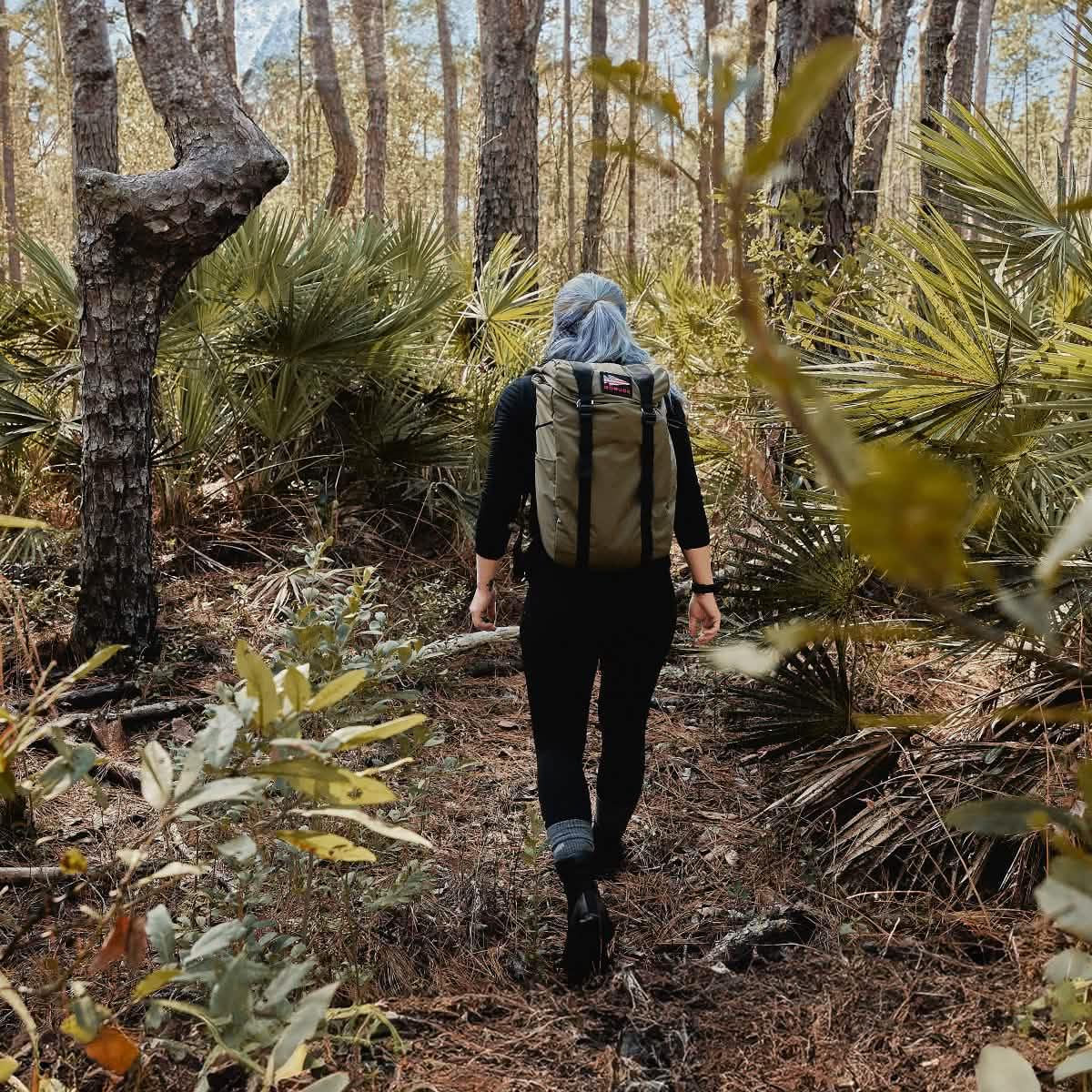 Wearing GORUCK’s Merino Challenge Socks, a hiker with a backpack treks through a lush forest surrounded by vibrant green foliage and tall trees.