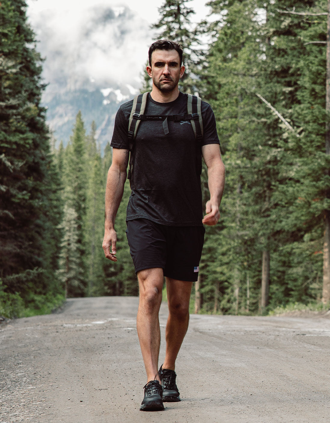 Man wearing black athletic clothes and backpack walks on a forest road with mountains in the background.