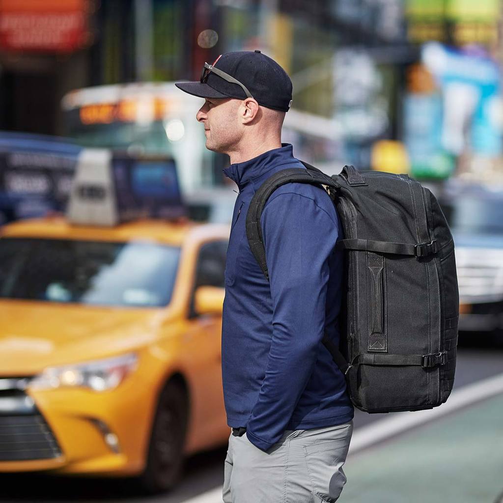 Man in a navy jacket and cap with a large black backpack stands by a yellow taxi on a city street.