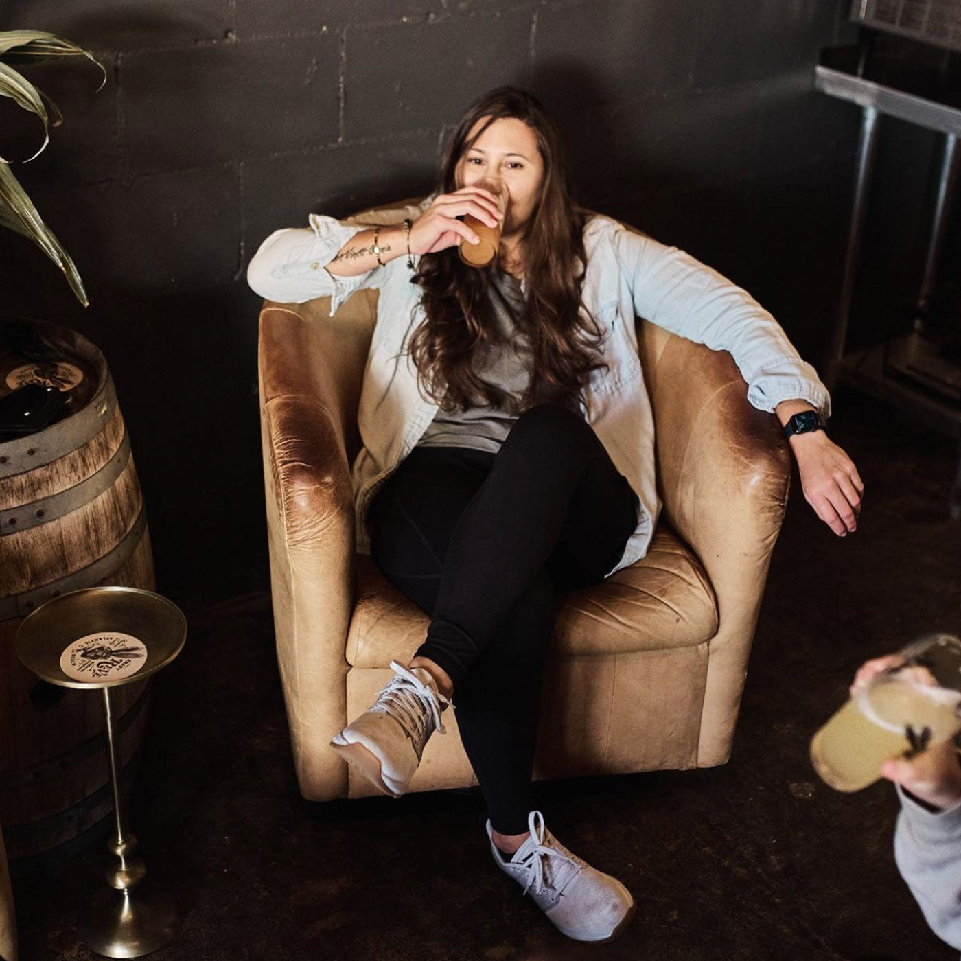 Woman sitting in a tan armchair, drinking from a glass, with one arm resting on the chair’s side.