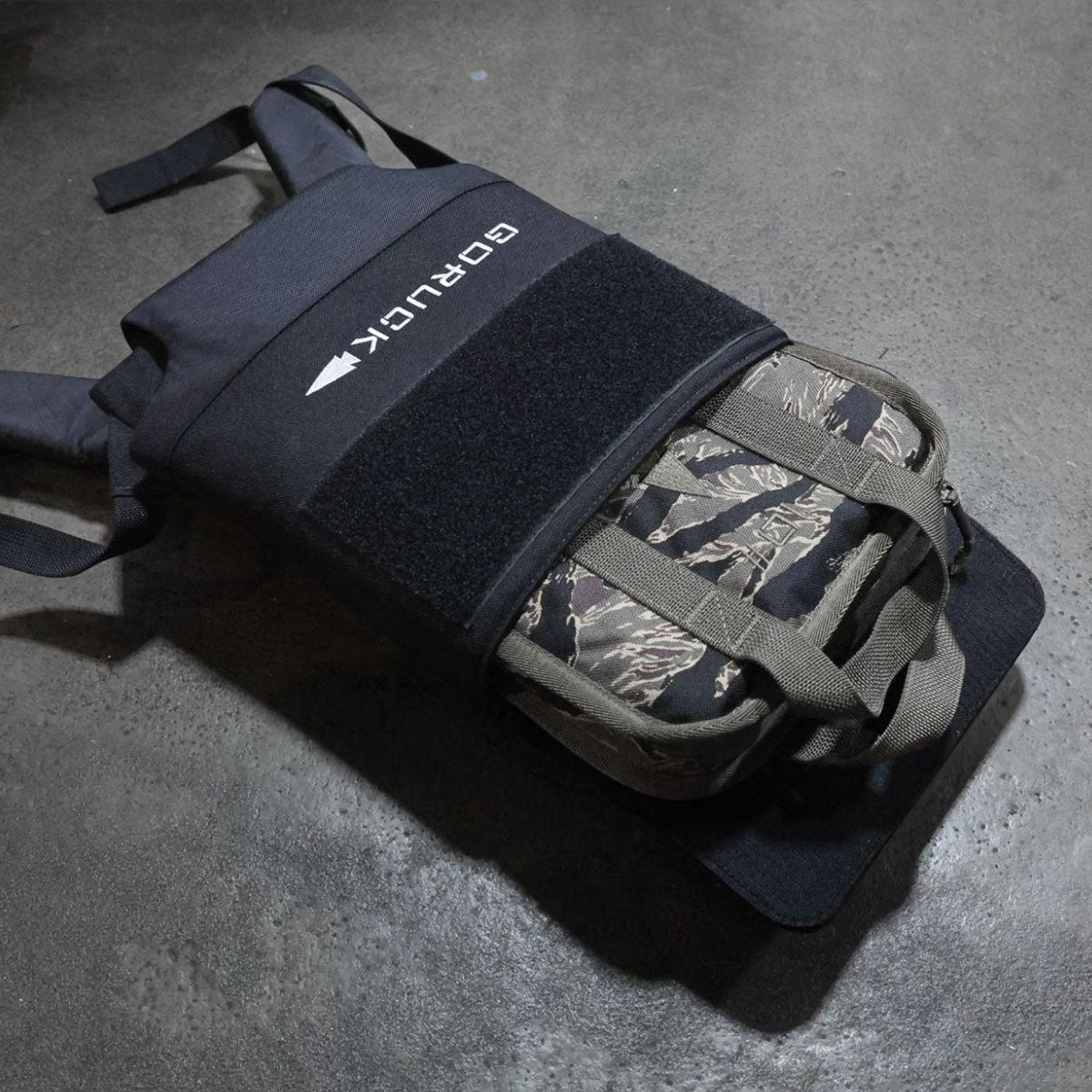 A black GORUCK backpack with a camouflage pouch attached, lying on a gray concrete floor.