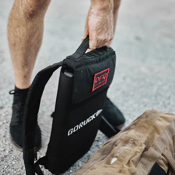 Person holding a black GORUCK backpack upright next to a tan bag on a gravel surface.