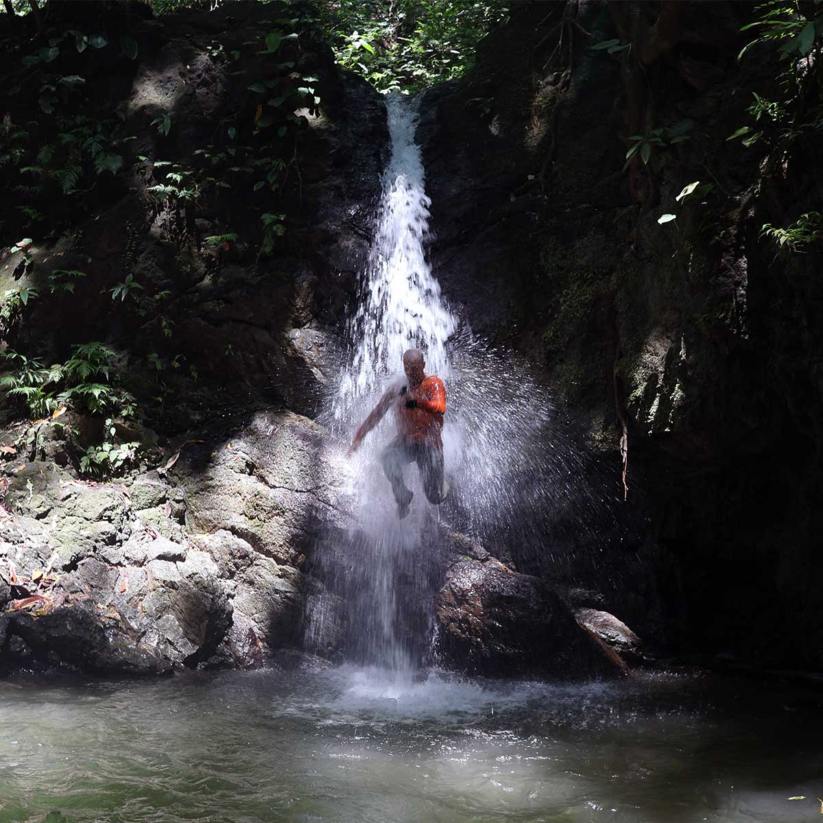 Person standing under a small waterfall surrounded by rocks and lush greenery.