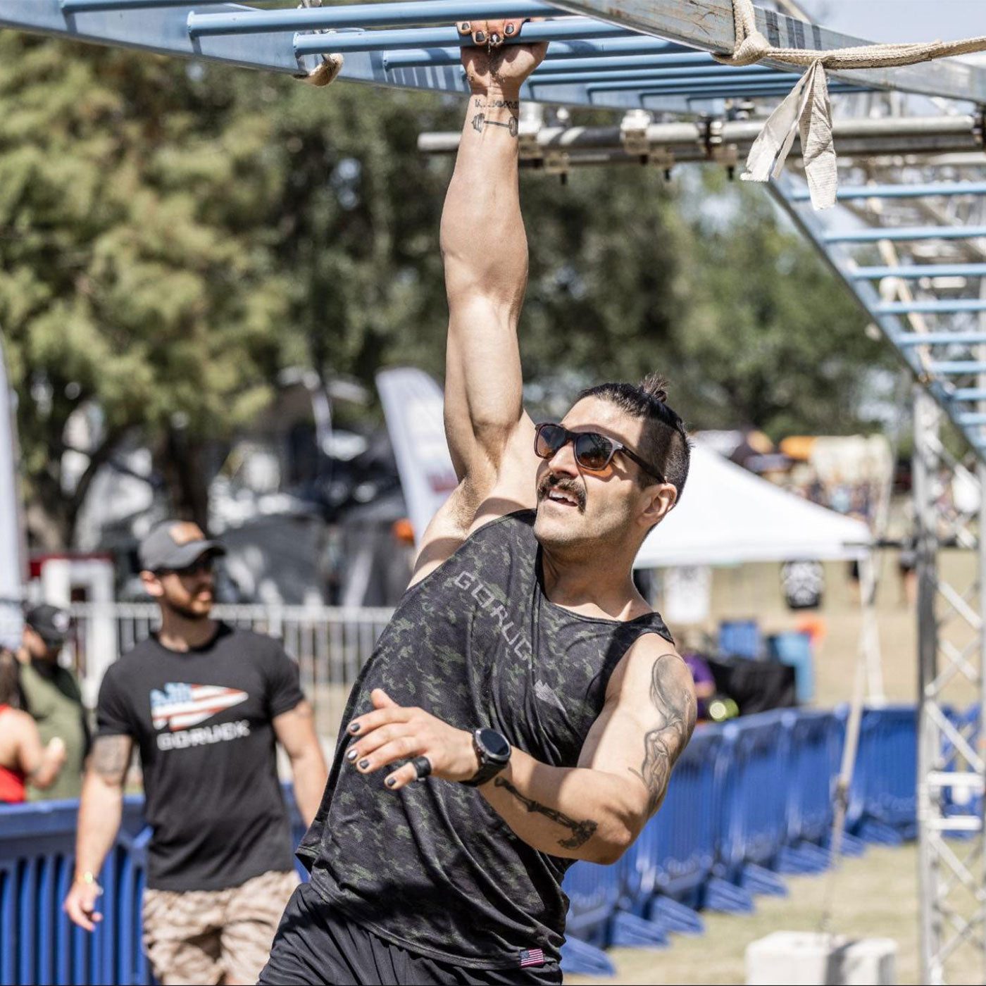 Man wearing sunglasses hangs from metal monkey bars during an outdoor obstacle course event.