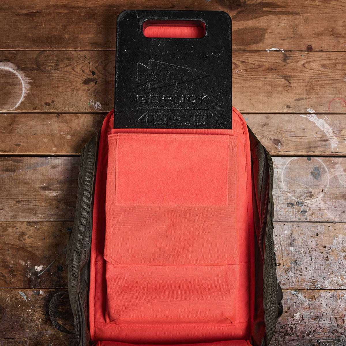 A 45 lb GORUCK ruck plate in an orange backpack on a worn wooden surface.