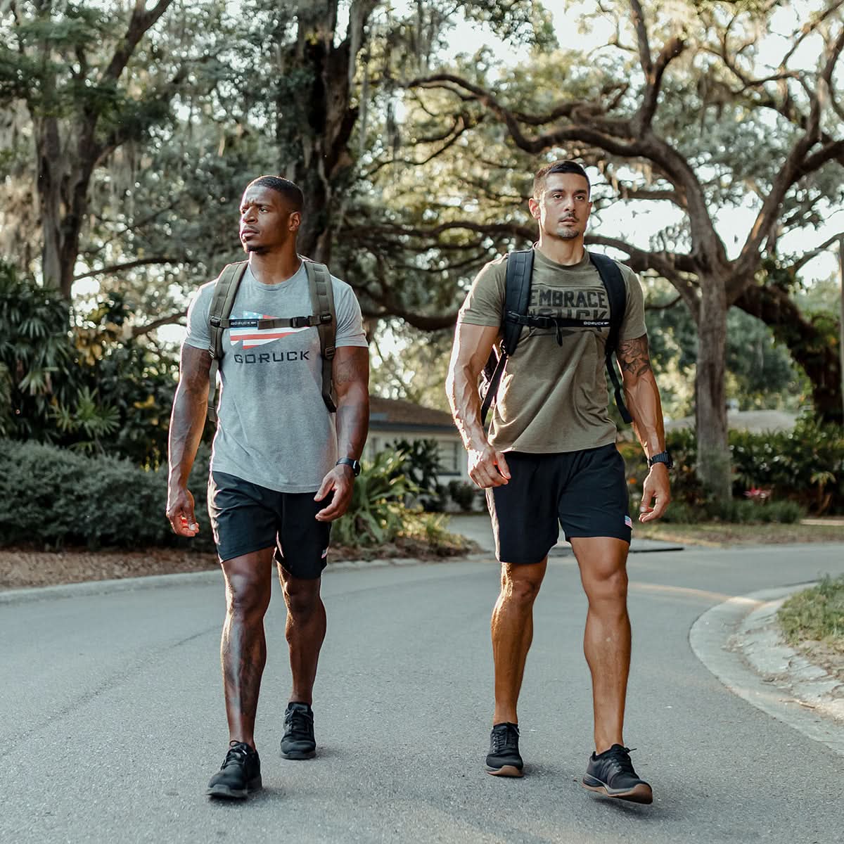 Clad in t-shirts and shorts, two men, who might be utilizing the GORUCK Ruck Clubs Starter Kit -Light, traverse a tree-lined road in an outdoor environment as they fully engage in the sport of rucking.