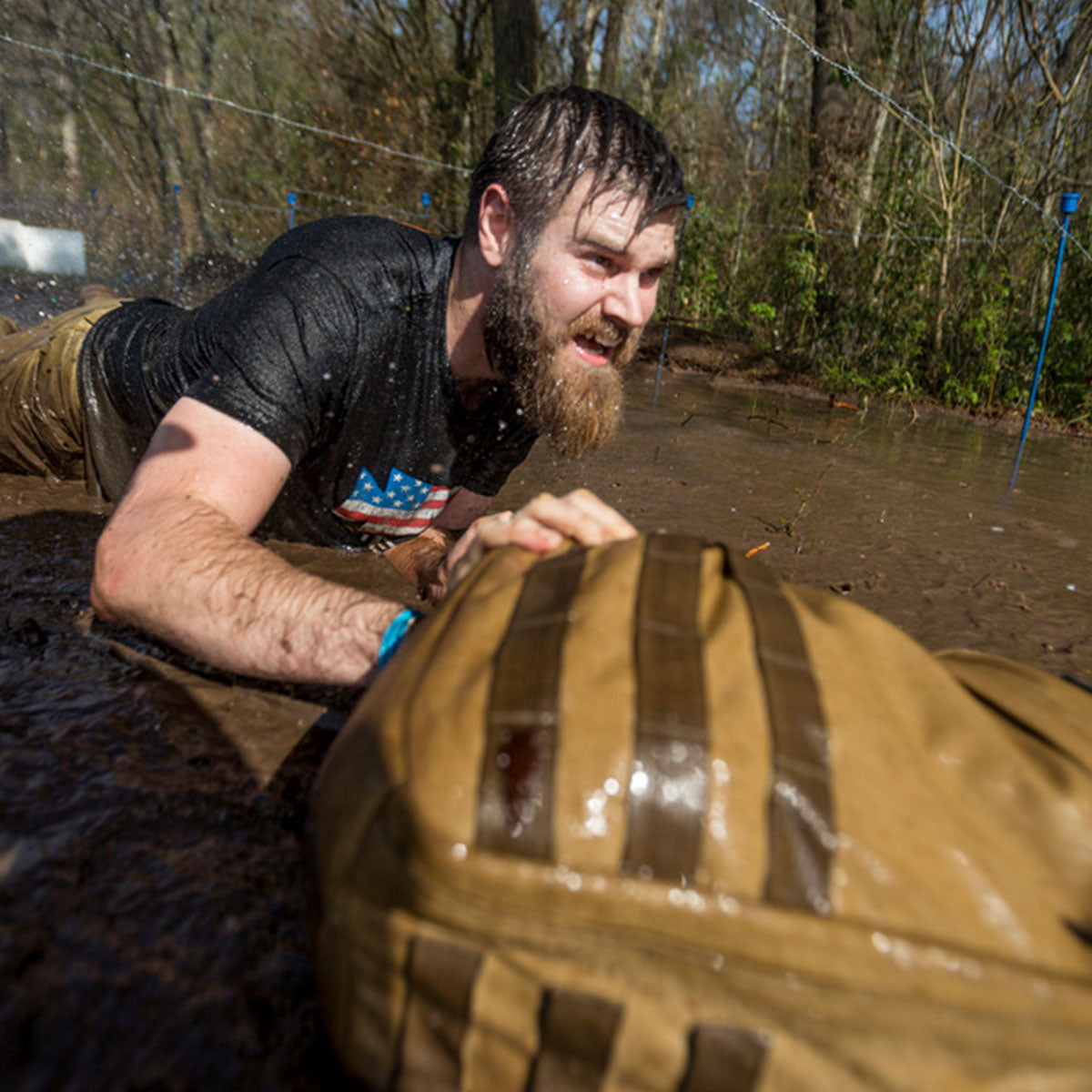 A bearded man crawls through muddy water outdoors, wearing a wet shirt and looking determined.