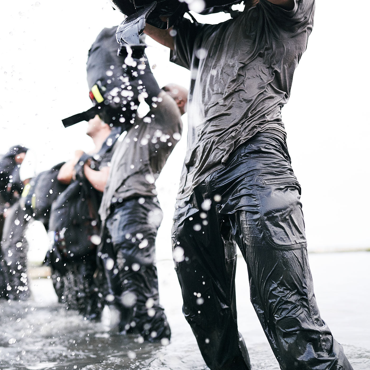 People in wet clothes lifting heavy bags above their heads, water splashing, standing in shallow water.