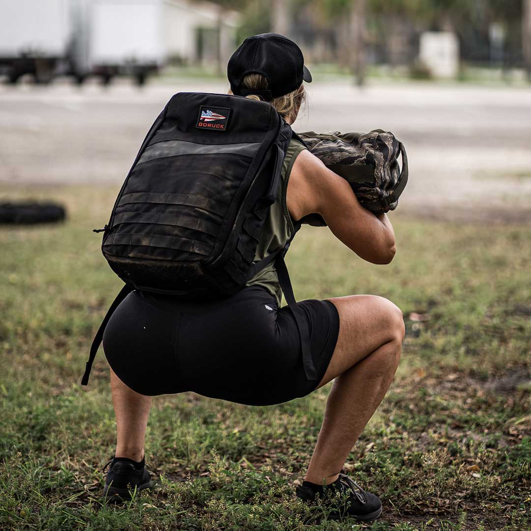 A woman in athletic wear squats outdoors, holding a sandbag with a large backpack on her back.