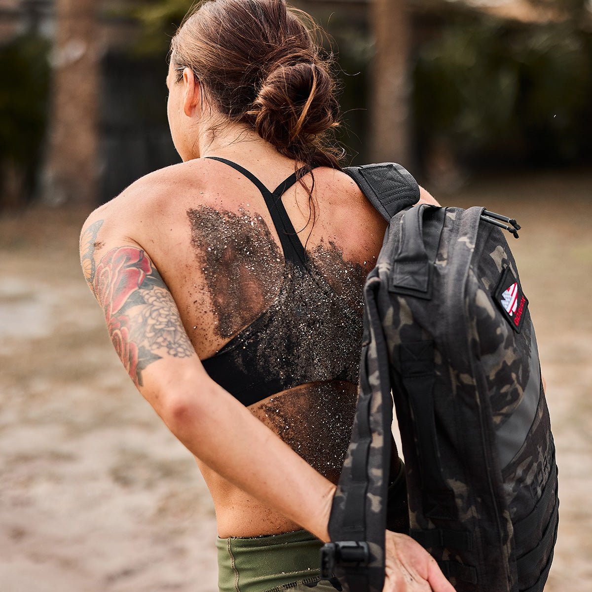 A woman with a tattoo and a dirty back carries a black backpack outdoors, wearing a sports bra and shorts.