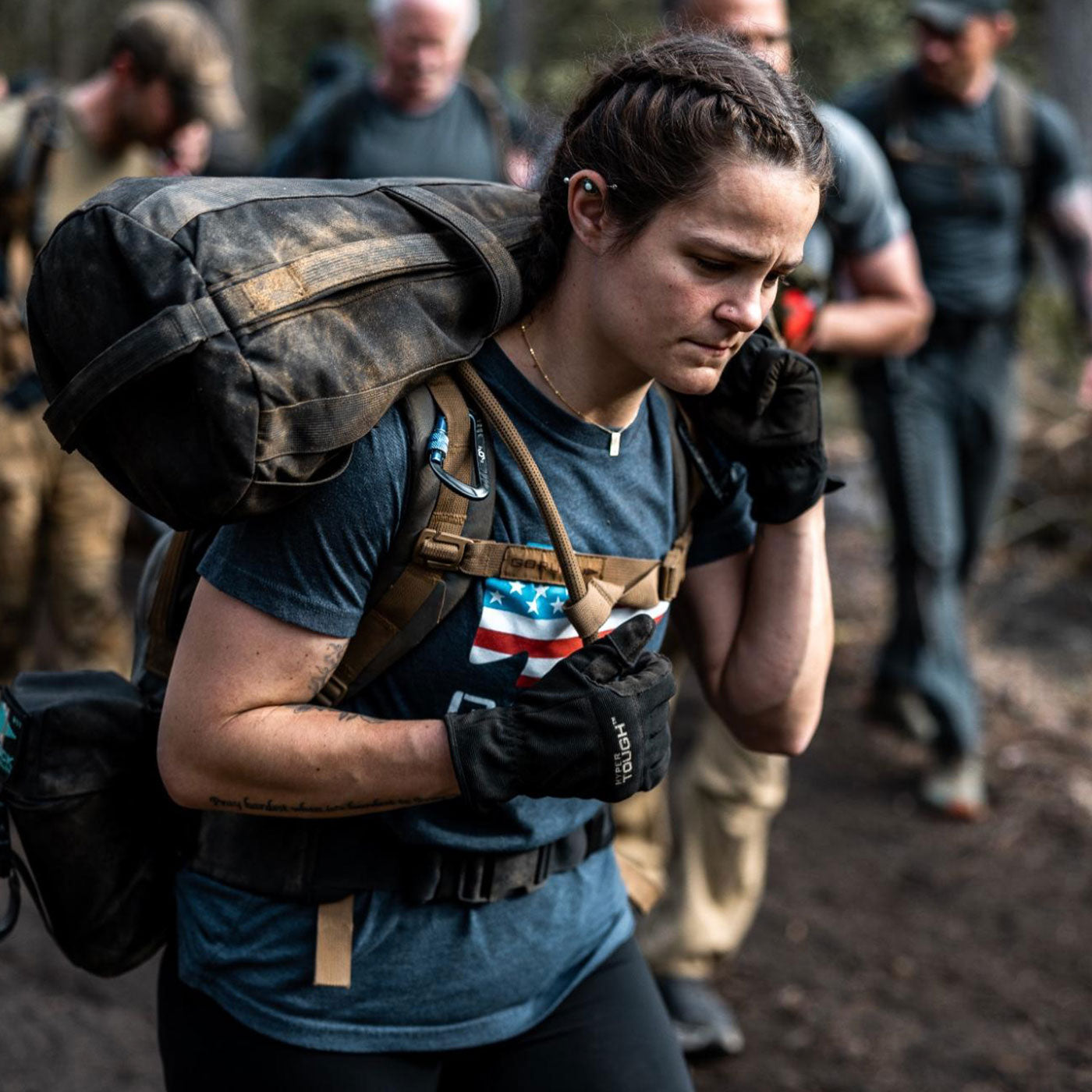 Woman with a large backpack hikes outdoors with a focused expression, surrounded by others in similar gear.