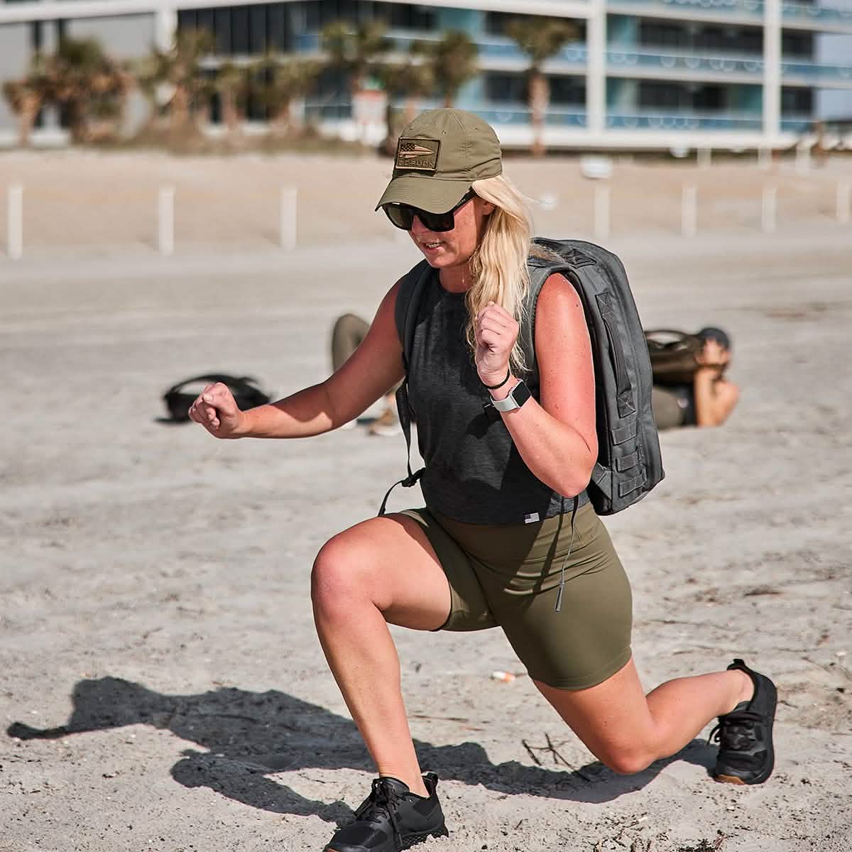 A woman wearing a cap, sunglasses, tank top, and GORUCK's Women’s Biker Shorts - ToughFlex lunges on a sandy beach carrying a backpack. In the background, another person exercises on the sand near modern buildings and palm trees.