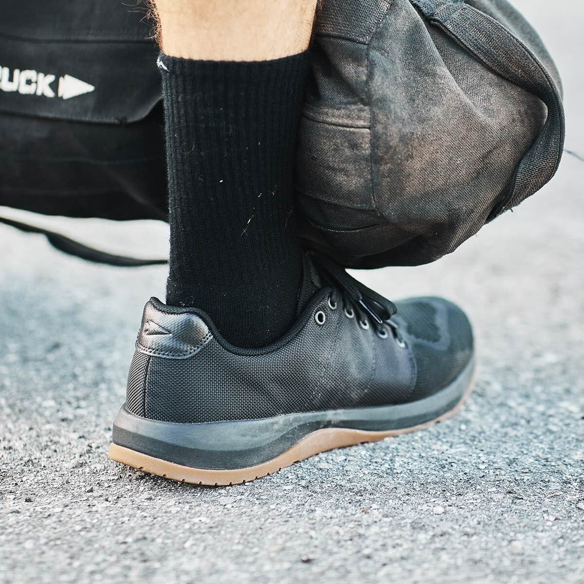 A person wearing black athletic shoes and socks rests a duffel bag on their foot on a paved surface.