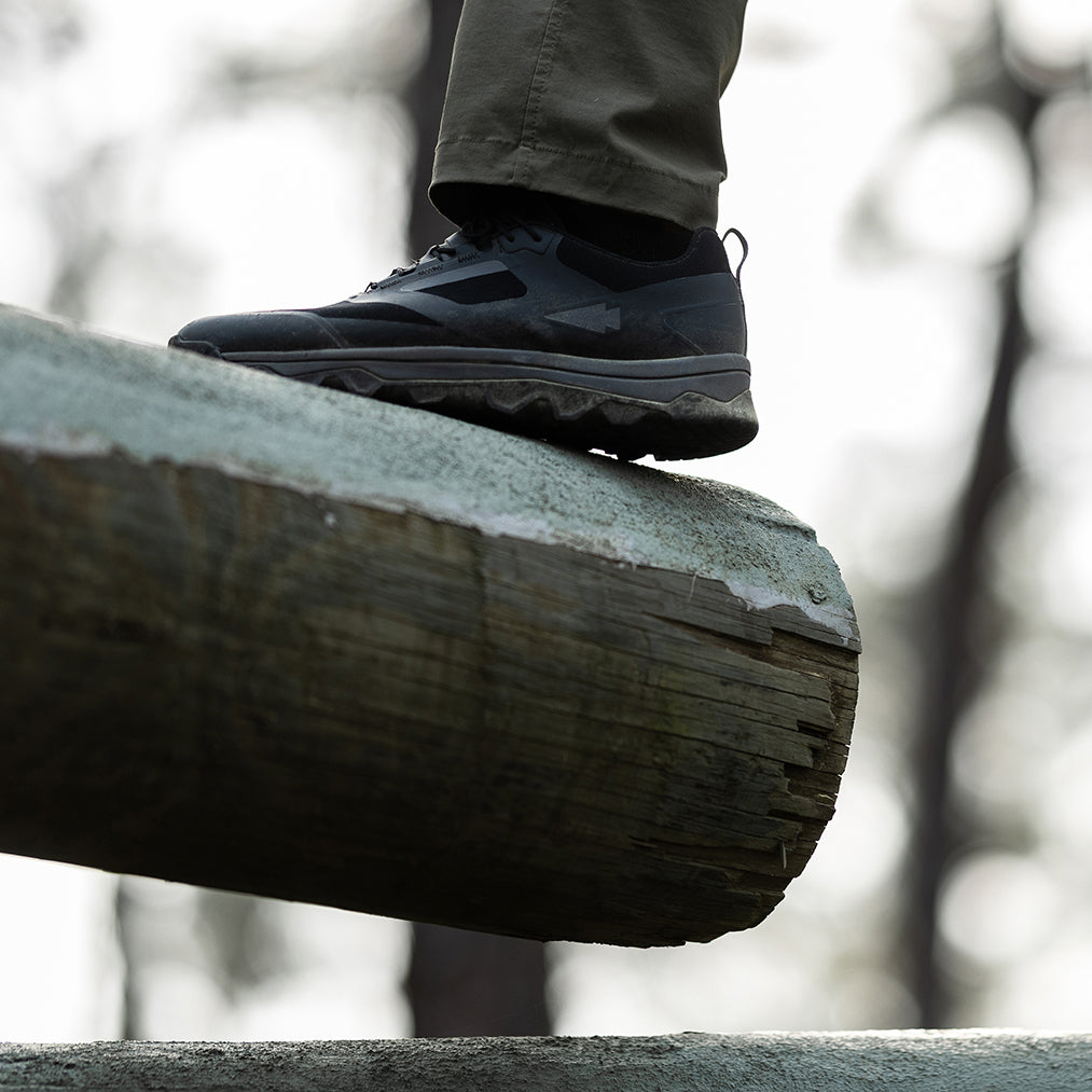 A person in dark sneakers and green pants balancing on a large, weathered wooden beam outdoors.