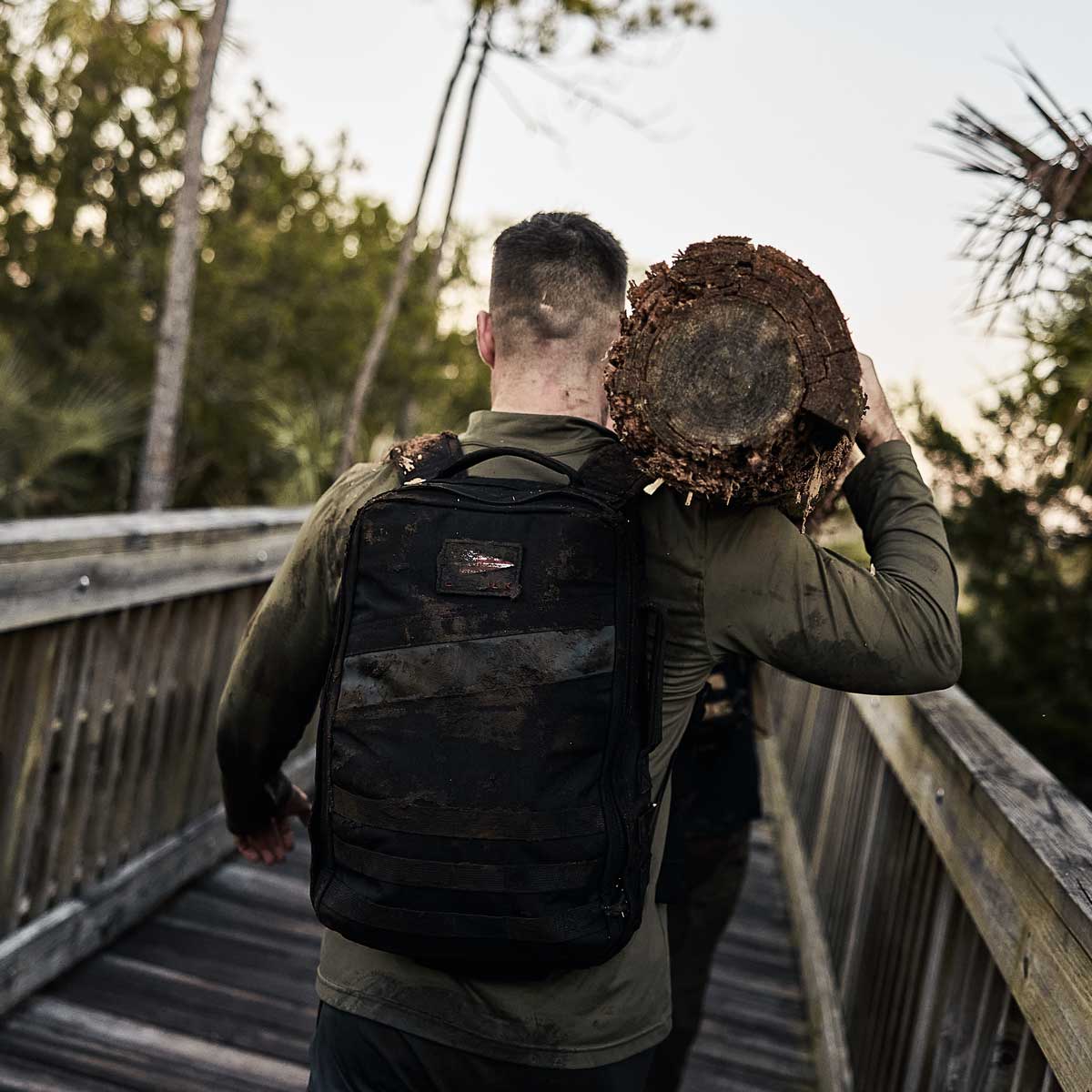 Man carrying a large log on his shoulder while walking on a wooden bridge in an outdoor setting.