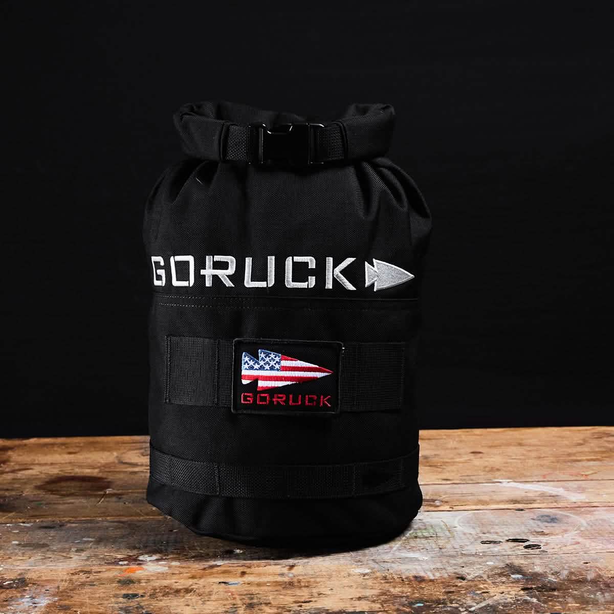 A black GORUCK Brick Bag (5-50 LBS) with an American flag patch rests on a wooden surface against a black background, showcasing its rugged design inspired by military gear.