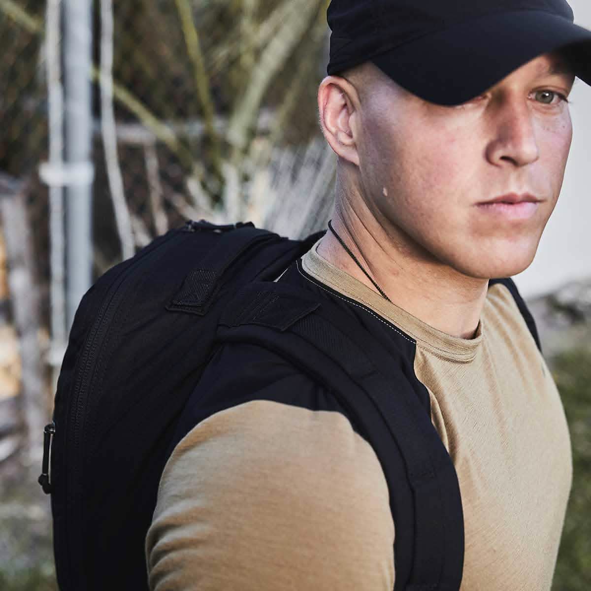A man outdoors near a fence wears a tan shirt, black cap, and carries a GORUCK Bullet Ruck Laptop backpack made of ballistic nylon Cordura with a bombproof laptop compartment.