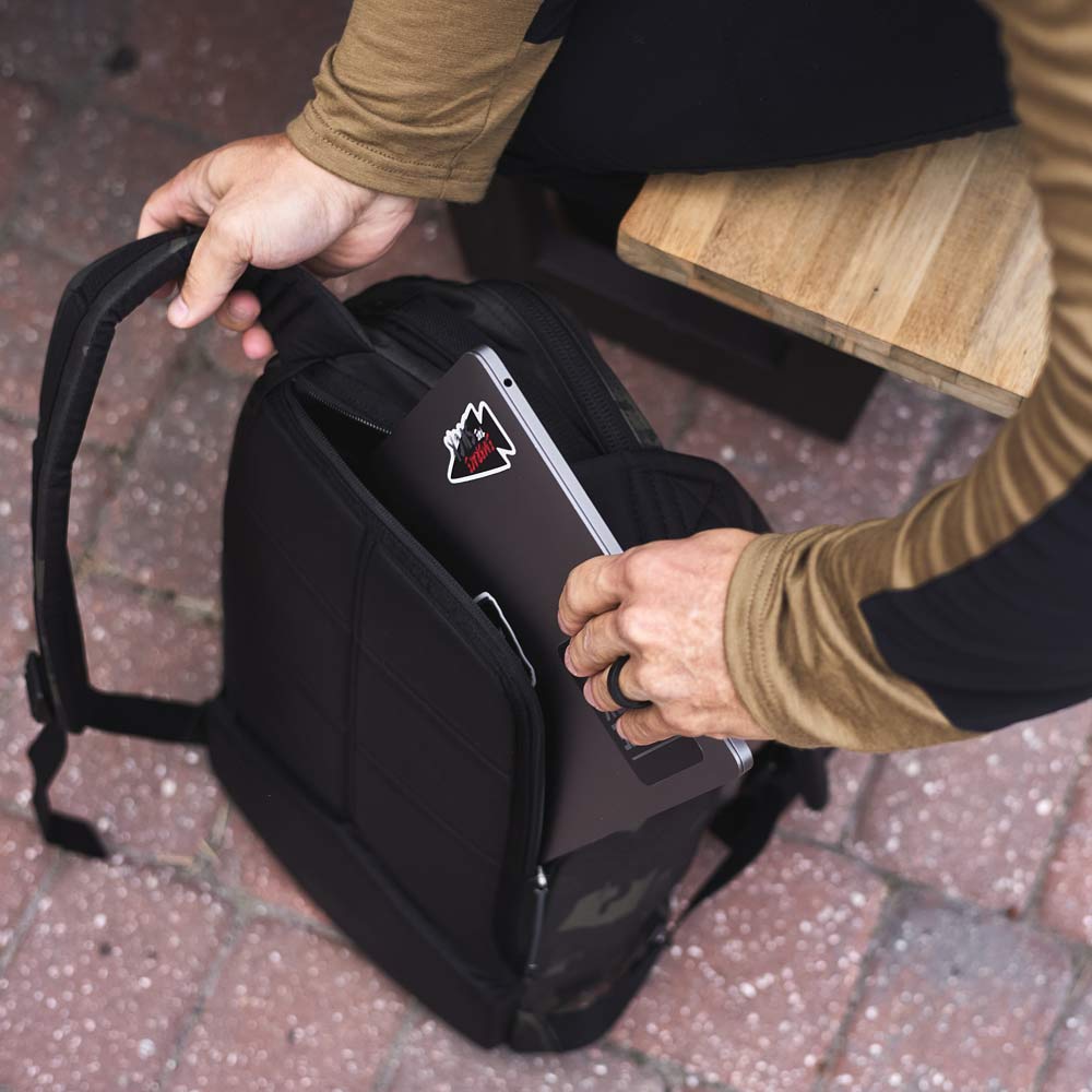 Person placing a laptop with stickers into a black backpack on a brick sidewalk next to a wooden bench.