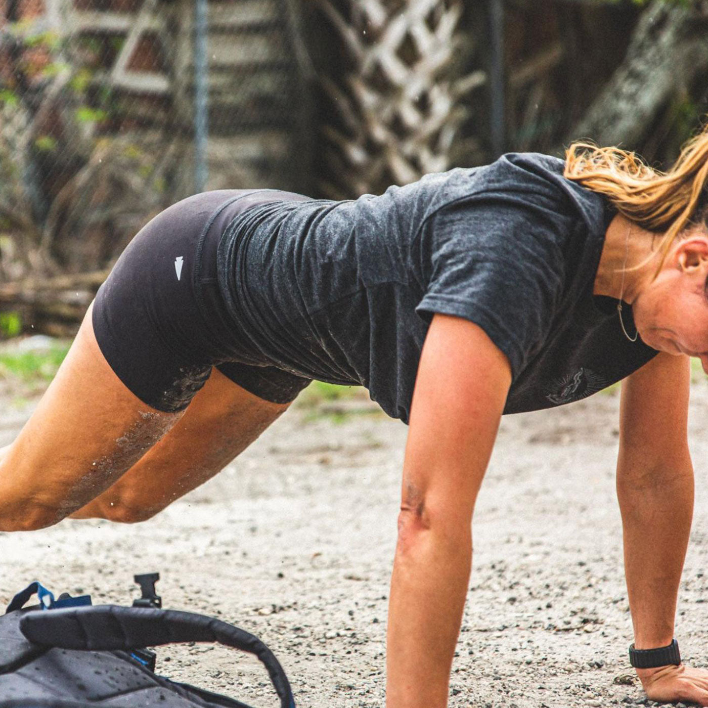 Woman outdoors doing a plank exercise on sandy ground, wearing athletic clothes, with a backpack nearby.