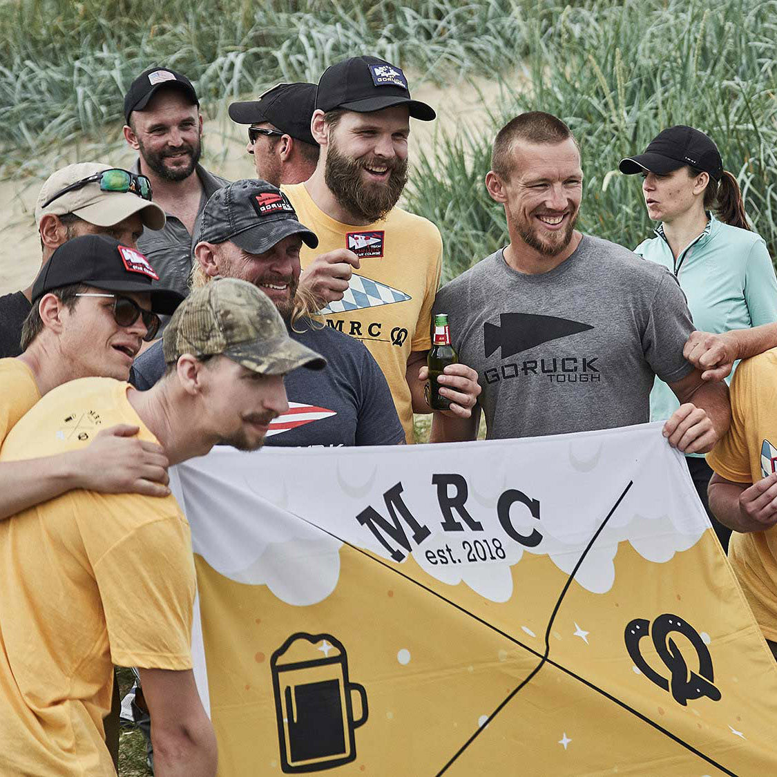 Group of smiling people outdoors holding a yellow MRC banner, with grassy dunes in the background.