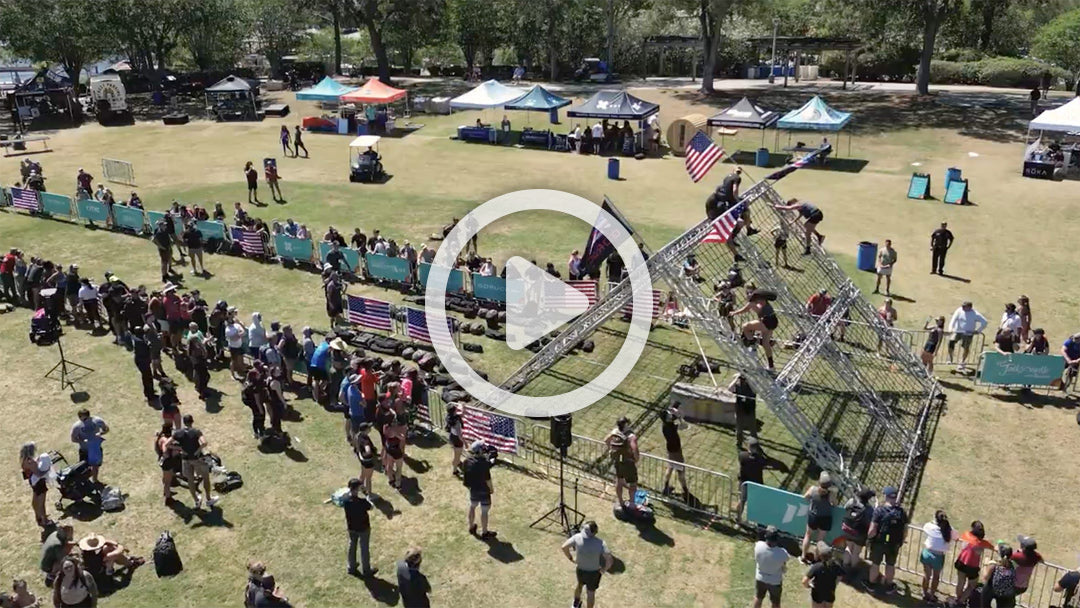 A crowd watches people climb a metal obstacle at an outdoor event with tents and flags.