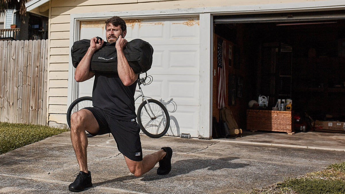 Man doing a lunge with a weighted bag on his shoulders outside a garage with a bicycle in the background.