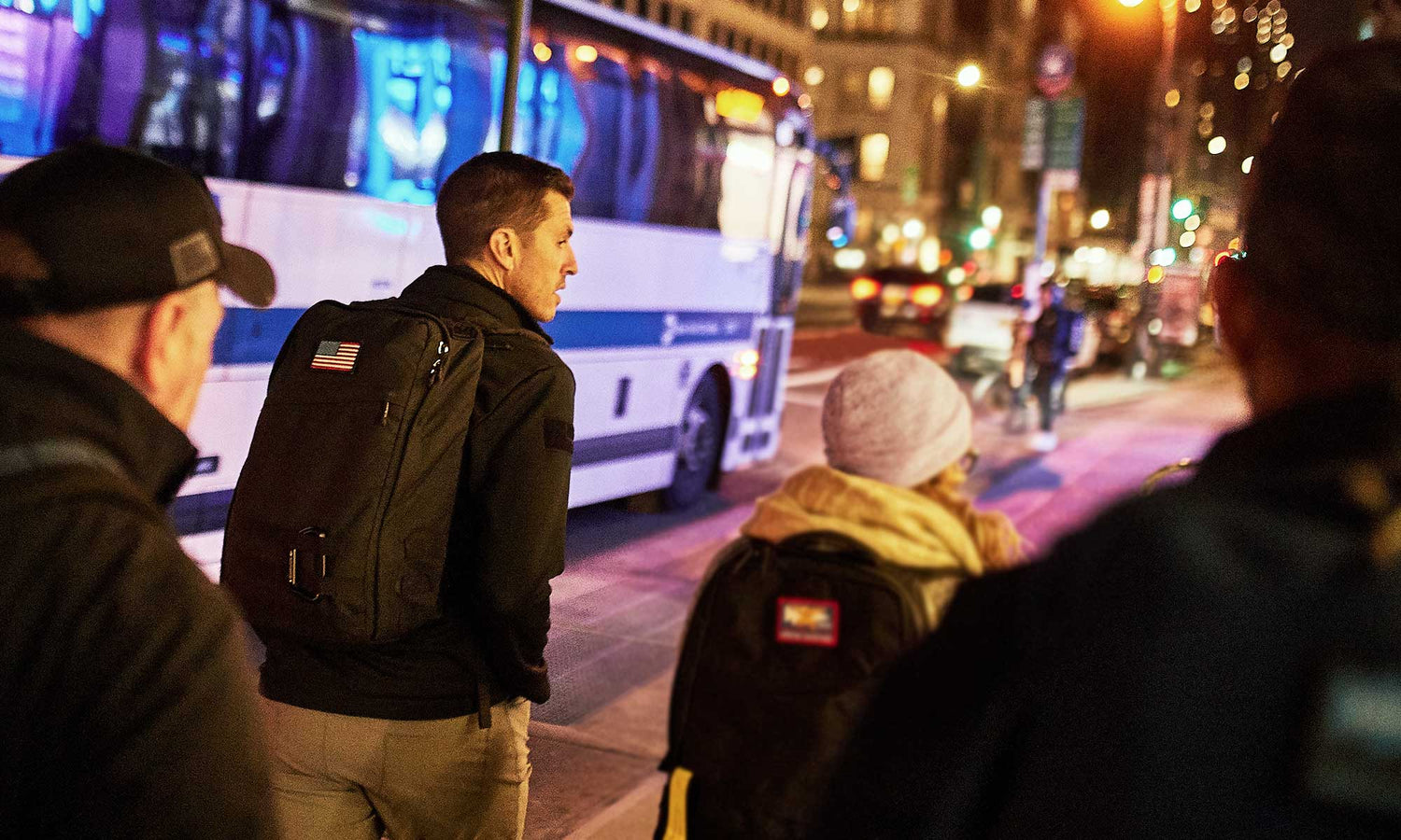 Four people with backpacks walk on a city sidewalk at night near a parked bus and bright street lights.