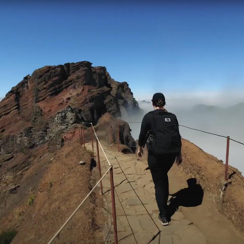 Person with a backpack walks along a mountain path with rocky peaks and clouds in the background.