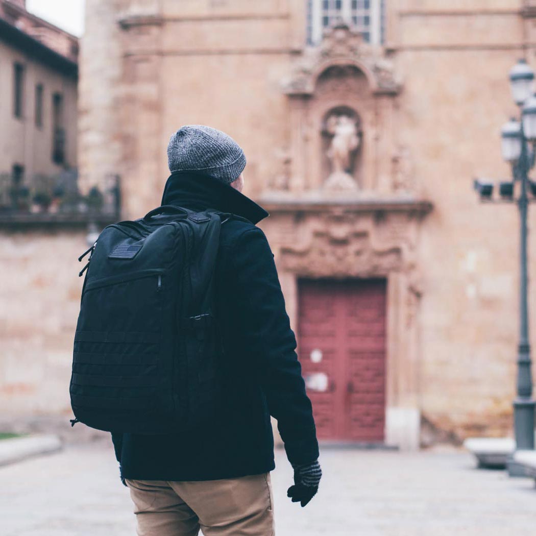 Person wearing a backpack and winter clothes stands in front of an old building with a red door.