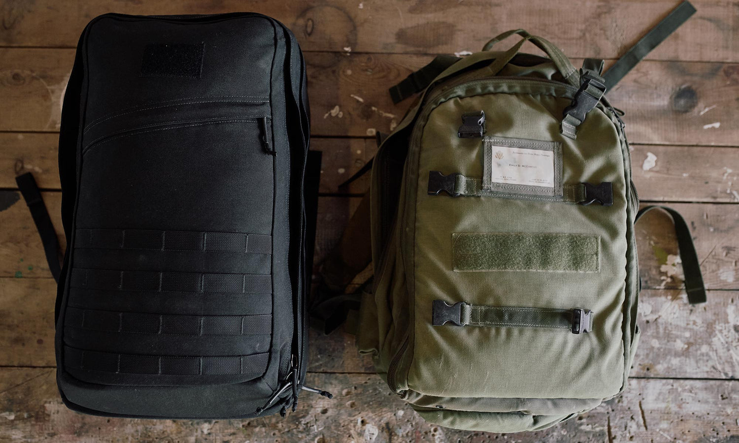 A black backpack and a green backpack placed side by side on a worn wooden floor.