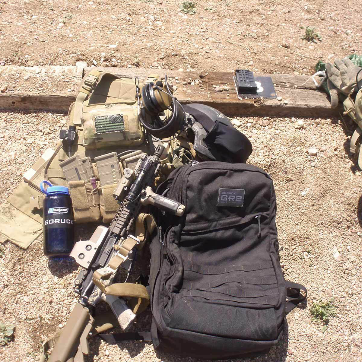 Tactical gear, rifle, backpack, headset, and water bottle laid out on gravel outdoors in sunlight.