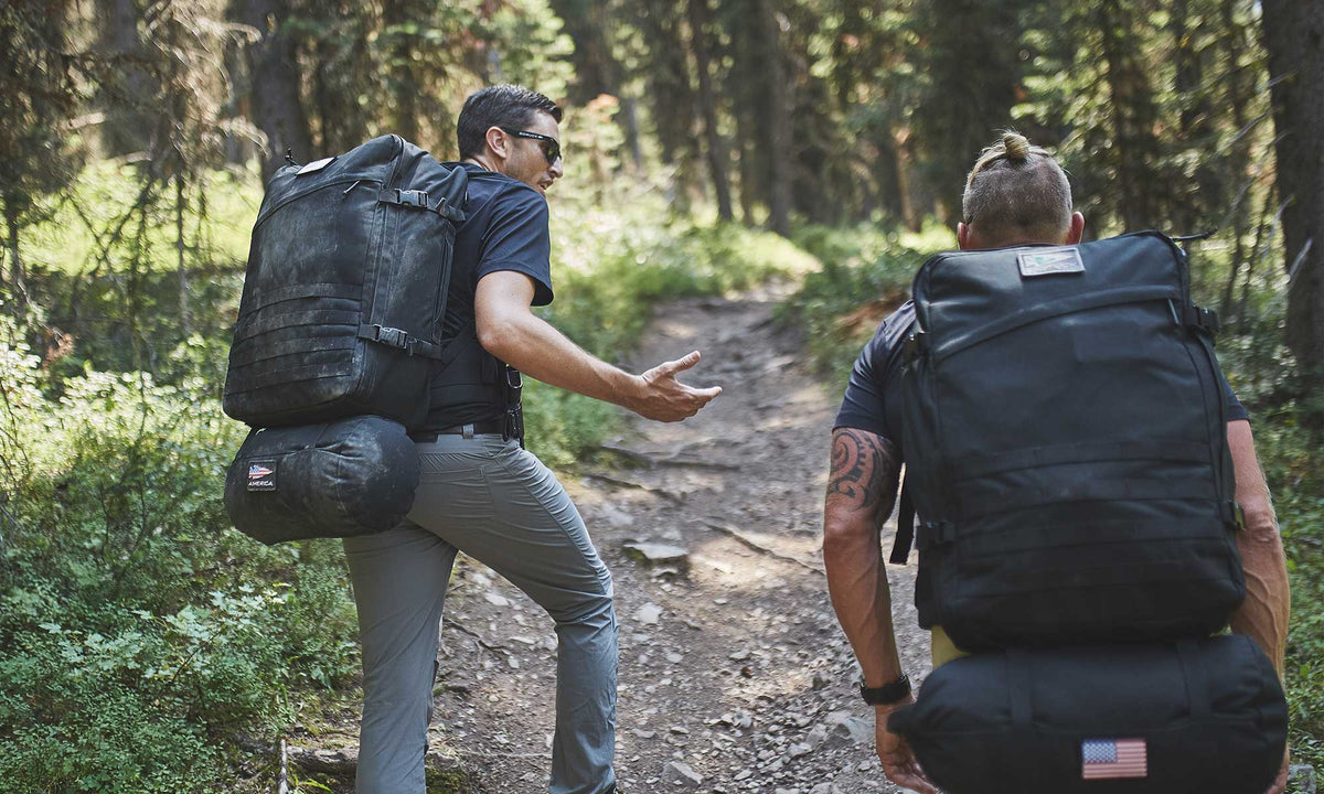 Two people with large backpacks hiking up a forest trail, talking and surrounded by trees and greenery.
