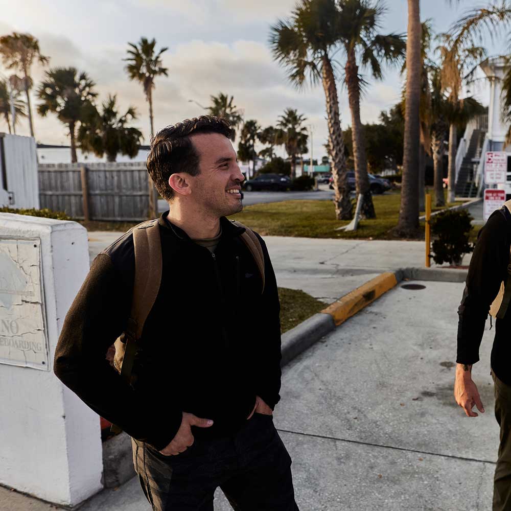 Man with a backpack smiling outdoors near palm trees and a parking area, with another person partly visible.