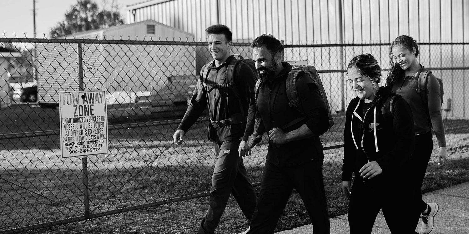 Four people wearing backpacks walk together, smiling, beside a chain-link fence in an outdoor area.
