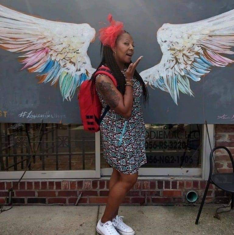 A woman poses playfully in front of a mural of large, colorful wings, making it look like she has wings.