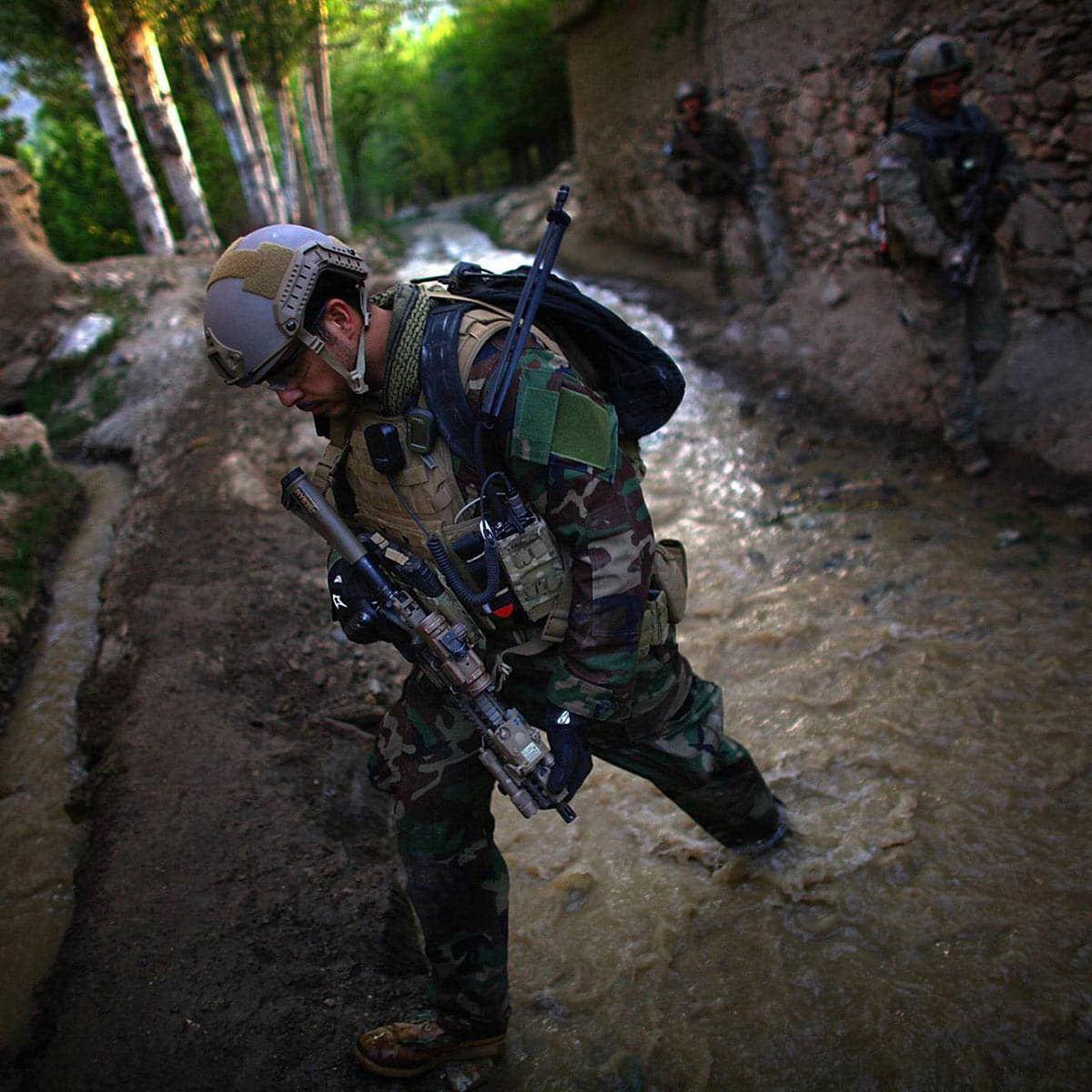 A soldier in camouflage crosses a shallow stream with rifle; two others follow in the background.